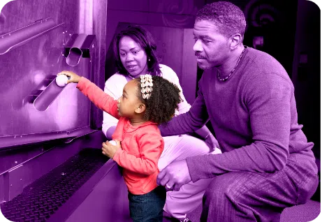 Parents (under a purple overlay) with their toddler (in full color) who is playing on an exhibit