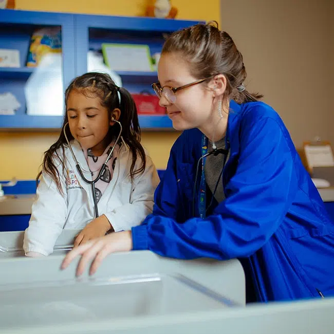 Child wearing a stethoscope and lab coat with an adult adn both are standing over an off camera subject