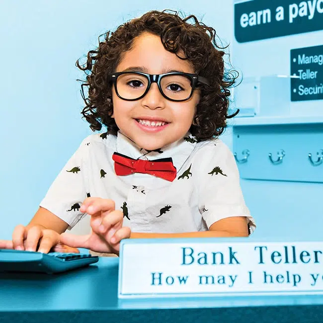 young boy wearing glasses, a red bowtie, and dinosaur collared shirt smiling at the camera and pretending to be a bank teller