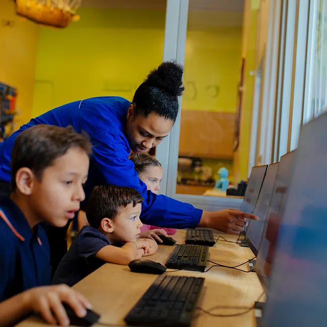 An adult volunteer standing over the shoulders of a child who is looking at a computer screen