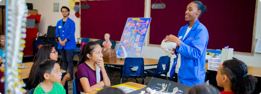 An adult docent wearing a Discovery Kids Museum blue labcoat presenting in front of children in a classroom setting