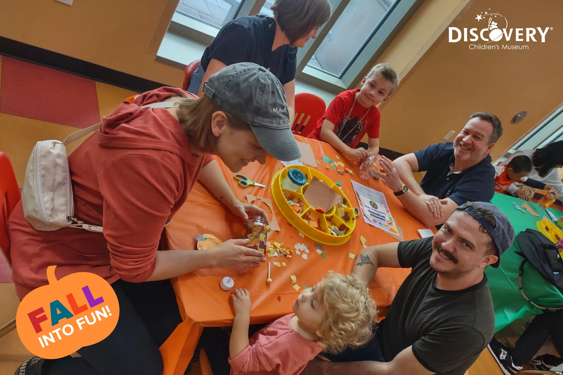 4 parents and 2 children sitting at small orange table with bits of colored paper at the Discovery Children's Museum