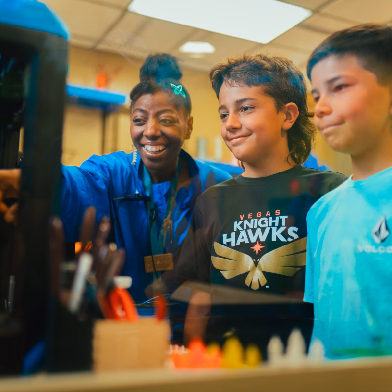"A Discovery Children's Museum staff member and two children watch a 3D printer in action during a summer camp session."