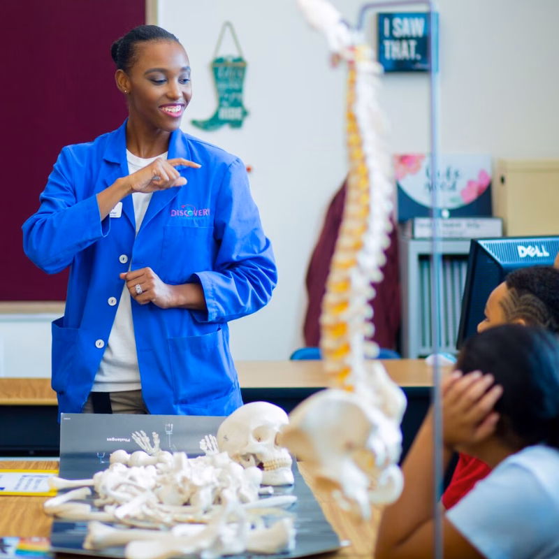 Educator Guide A DISCOVERY Children's Museum educator in a blue lab coat presenting a human skeleton model and bone specimens to students during an outreach program.